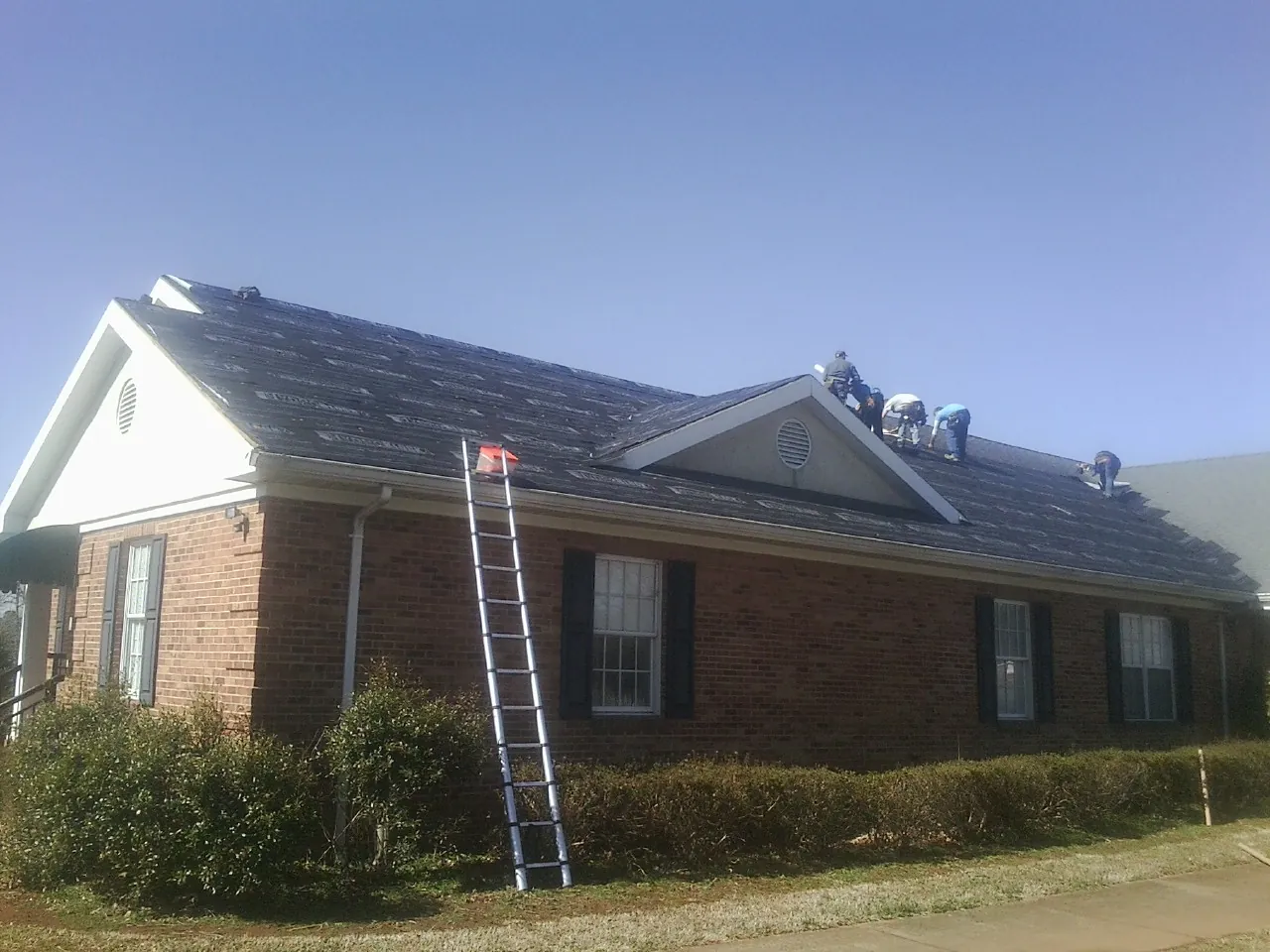 Crew tearing shingles off a brick ranch roof with a ladder set — Metal Roof Repair work by our crew in Little Switzerland