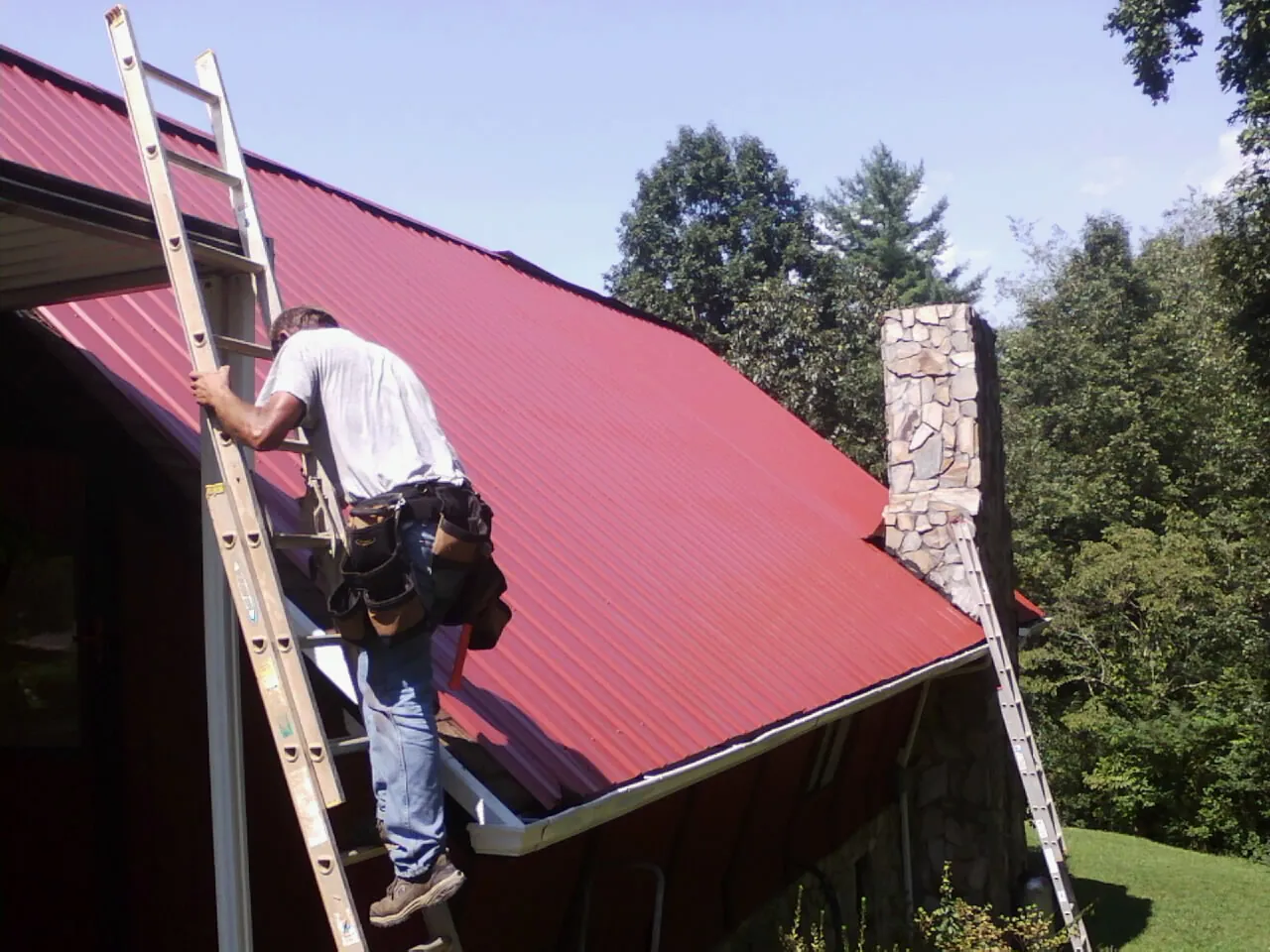 Roofer on a ladder beside a red metal roof and stone chimney — Soffit & Fascia Repair by Roofing Brothers in Little Switzerland